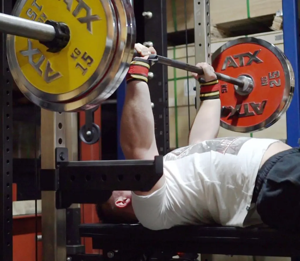 Person performing a bench press with a barbell in a gym. The barbell has red and yellow weight plates marked with ATX. The individual is lying on a bench, wearing wrist wraps and a white shirt.