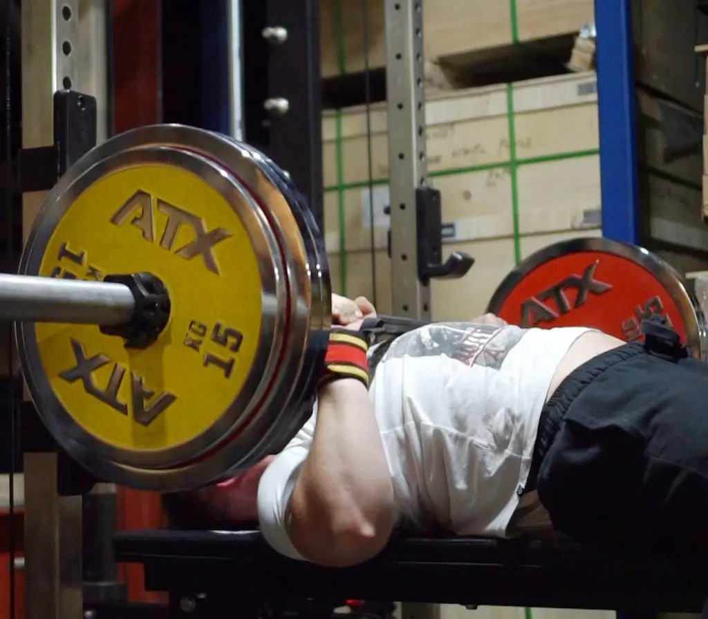 A person is performing a bench press in a gym. The barbell is loaded with large yellow and red weight plates labeled ATX. The individual is lying on a bench, focusing on lifting the weight. Boxes and shelving are visible in the background.
