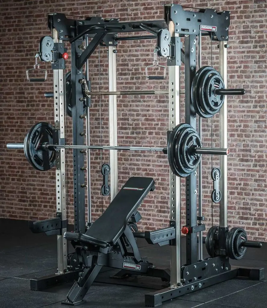 A weightlifting station with a barbell loaded with weights on a squat rack and an adjustable bench. The setup is positioned on a dark gym floor with a brick wall backdrop. Various handles and attachments are visible.