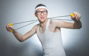 A person wearing glasses, a white headband, and a white tank top is holding a resistance band with yellow handles, stretching it with a playful expression against a gray background.
