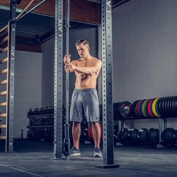 A shirtless man in gray shorts exercises with determination on a cable machine at a gym, equipped with kettlebells, weight plates, and a wooden ladder, alongside an ATX® Barbell Storage system.