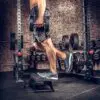 In front of brick walls and gym equipment, a person does step-ups on a low platform surrounded by racks of dumbbells. Holding two ATX® Calf Blocks, they showcase their strength and focus, dressed in athletic apparel and shoes.