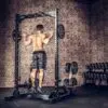 A person performs pull-ups on a metal frame in a brick-walled gym, wearing shorts and sneakers. Nearby is an ATX® Calf Block, while weight plates are stored on racks in the background.