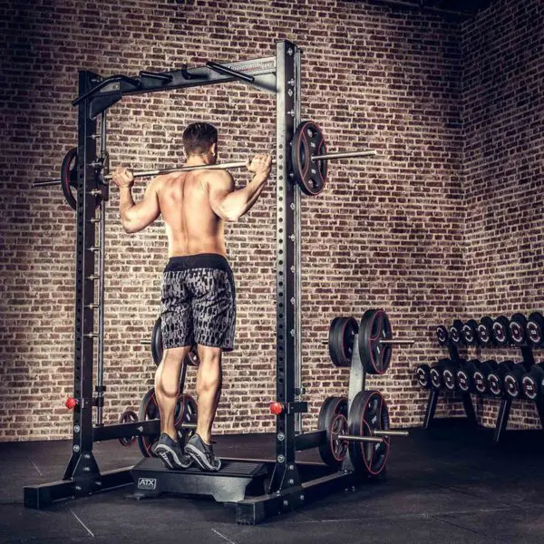 A person performs pull-ups on a metal frame in a brick-walled gym, wearing shorts and sneakers. Nearby is an ATX® Calf Block, while weight plates are stored on racks in the background.