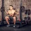 A shirtless man squats with a barbell, using an ATX® Calf Block for added intensity, against a brick wall in the gym. Weight plates are stacked beside him on the rack.