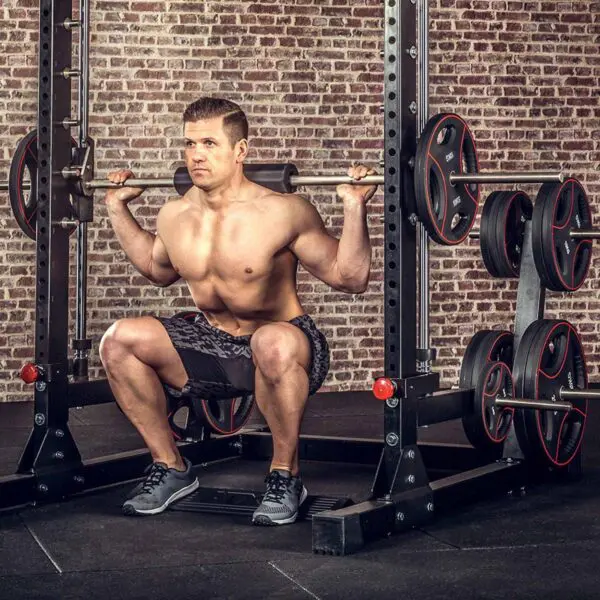 A shirtless man squats with a barbell, using an ATX® Calf Block for added intensity, against a brick wall in the gym. Weight plates are stacked beside him on the rack.