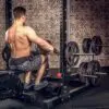 A person in shorts is seated on a gym bench performing a cable row, with the ATX® Calf Block complementing the equipment, including a weight rack with multiple plates, set against a brick wall backdrop.