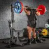 A person in black athletic clothing performs a squat with a heavily loaded barbell on an ATX® Competition Combo Rack in a gym. Weight plates are stacked on the floor nearby against a brick wall background.