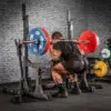 A person squats with a loaded barbell on their shoulders using the ATX® Competition Combo Rack. The barbell has red and blue weight plates, set against a gym backdrop featuring equipment and a brick wall.