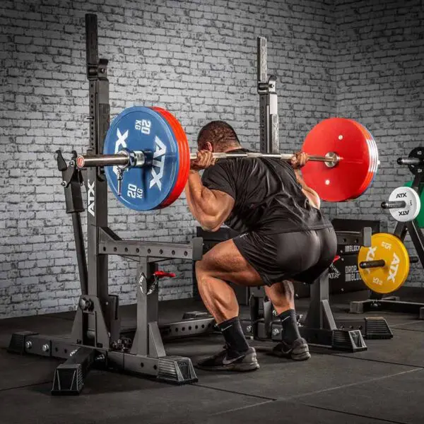 A person squats with a loaded barbell on their shoulders using the ATX® Competition Combo Rack. The barbell has red and blue weight plates, set against a gym backdrop featuring equipment and a brick wall.