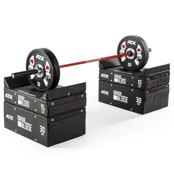 A barbell with weights is balanced on stackable black ATX® 30 cm Soft Drop Blocks labeled 15 cm and 30 cm. The weights are marked ATX with numbers, indicating a setup for weightlifting or strength training exercises.