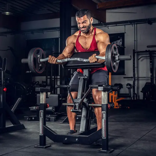 A determined man in a red tank top and black shorts performs an intense workout with the ATX® EZ Bar Curl Camo - DISCONTINUED amid various gym equipment.