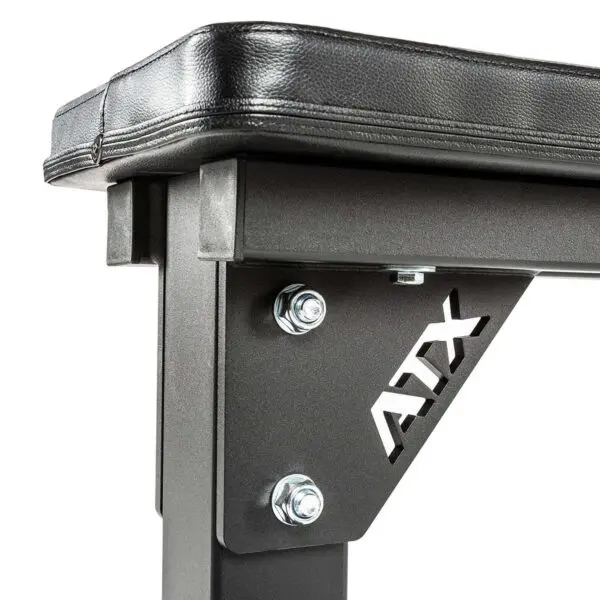 A close-up reveals the ATX® Flat Bench, showcasing its sturdy black frame and cushioned seat. Metal bolts secure the structure, and a cut-out ATX logo graces the side support, emphasizing durability and strength for heavy-duty workouts.