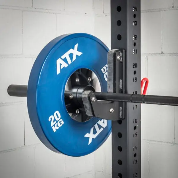 An ATX® J-Hooks Type III holds a 20 kg blue weight plate on a barbell in a squat rack against a white brick wall with numbered markings.