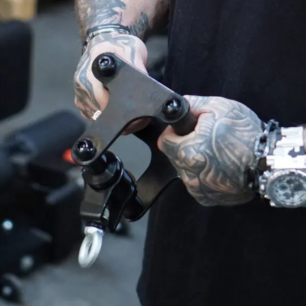 In a bustling workshop, a person with tattooed arms and a bulky watch holds the ATX® Lat Bar Blackline, featuring multiple bolts and an end hook.
