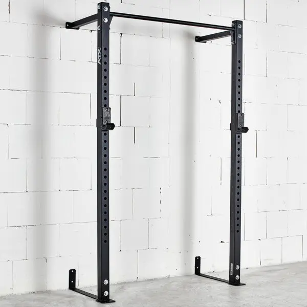An ATX® Wall Mounted Squat Rack, black and featuring a pull-up bar, is installed on a white brick wall. It has adjustable J-hooks for securing a barbell. The concrete floor is clear of weights and other equipment.