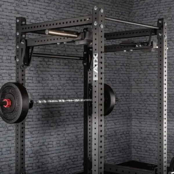 A black ATX® Jammer Arms power rack is set against a gray brick wall, featuring a loaded barbell with black weight plates. The rack includes multiple adjustment holes, displaying the ATX brand name on the side.