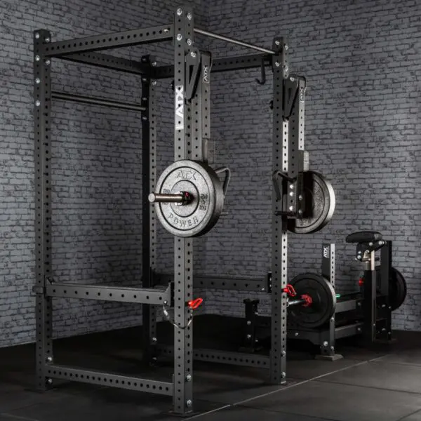 A gym featuring ATX® Jammer Arms attached to a power rack holds a barbell with weight plates against a textured gray brick wall. The floor is covered in black mats, and an incline bench press with extra weights is visible in the background.