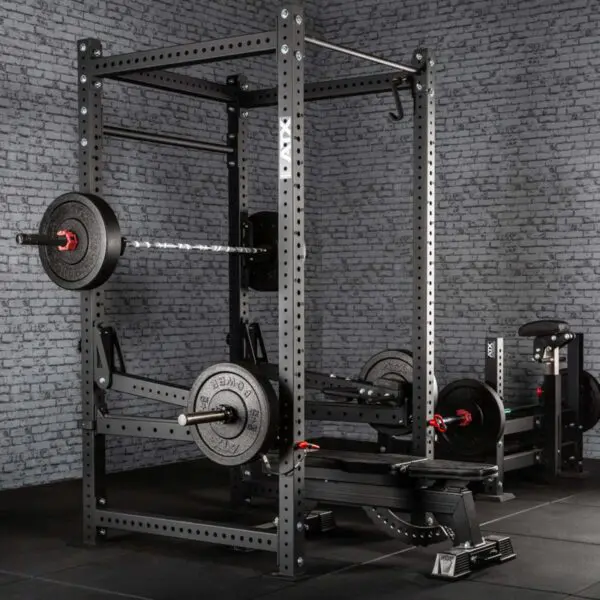 A home gym setup featuring ATX® Jammer Arms with a power rack, barbell, and weights on a dark floor against a gray brick wall. Additional fitness machines and weight plates can be seen in the background.