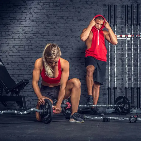 In a gym, a woman wearing a red sports top and black shorts kneels to adjust the weights on an ATX® Olympic Camo Power Bar. A man in a red sleeveless hoodie stands nearby, fixing his hood amid various gym equipment.