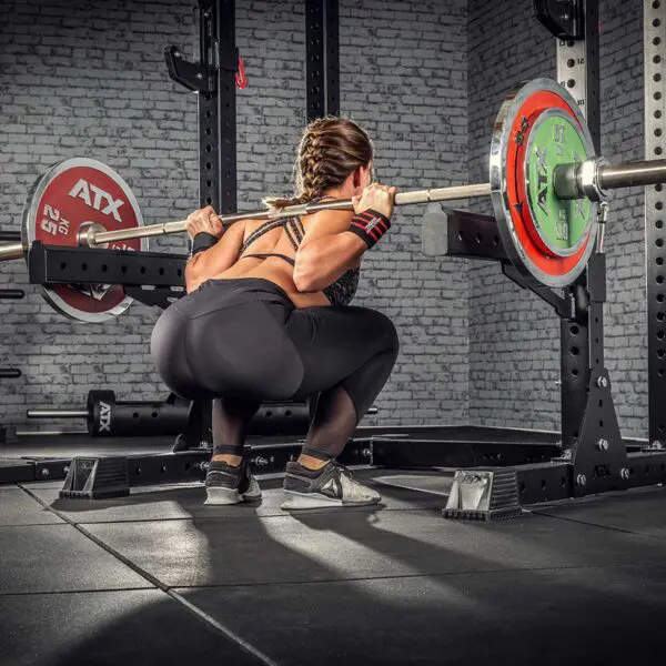 Using the ATX® Competition Powerlifting Barbell, a woman dressed in athletic gear focuses on her form and strength as she performs a weighted squat in the gyms squat rack.
