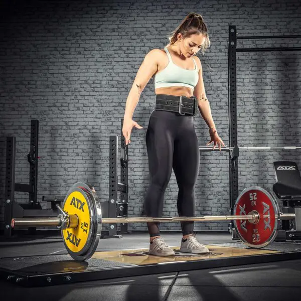 A woman stands ready to lift an ATX® Competition Powerlifting Barbell at the gym, wearing a white sports bra, black leggings, a lifting belt, and sneakers. Behind her are a brick wall and various workout equipment.