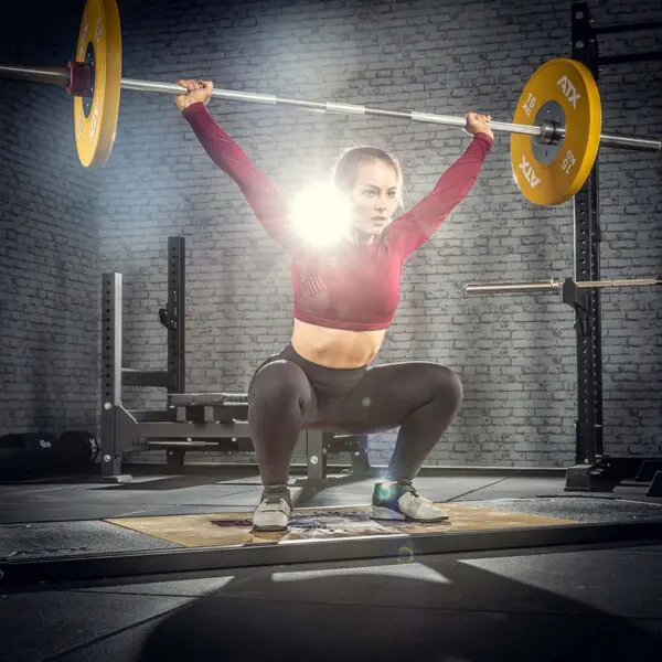 An athlete in Olympic weightlifting attire performs an overhead squat with the ATX® Competition Olympic Weightlifting Barbell in a well-equipped gym, under bright lights that enhance focus and strength.
