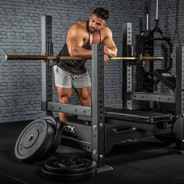 A man in a gym prepares a bench press by adjusting an ATX® Olympic Golden Powerlifting Bar on a squat rack. He wears a tank top and shorts, with weight plates on the floor nearby and brick walls visible in the background.