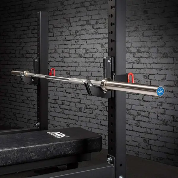 The ATX® Olympic Stainless Steel Bar is on a squat rack with safety hooks and a padded bench below, set against a textured gray brick wall, creating a gym environment.