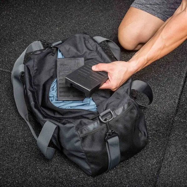 On a textured rubber floor, a person in gray athletic shorts kneels next to their trusty Mini Squat Wedges. They place a heavy weight plate into a black gym bag with teal interior lining and a wide strap for easy carrying.