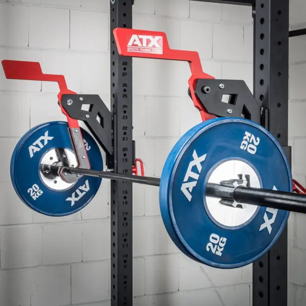 In a gym, an ATX® Mono Lift secures a weightlifting bar with blue 20 kg plates on a wall-mounted bracket. The scene includes a white brick wall and part of a metal rack in the background.