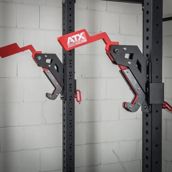 Two ATX® Mono Lift adjustable safety spotters, in red and black with ATX on the levers, are attached to a power rack against a white brick wall.