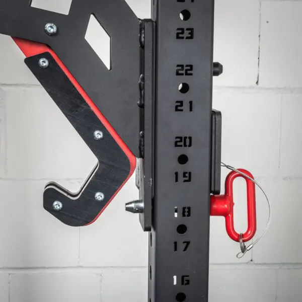 A close-up of the ATX® Mono Lift, a black metal squat rack with numbered height adjustments and a mounted J-hook, featuring red accents. The equipment is set against a white brick wall.