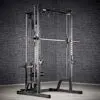 A black ATX® Smith Machine System Plate Loaded is set on a gray floor against a dark brick wall, featuring a pull-up bar, adjustable safety bars, and weight storage pegs.