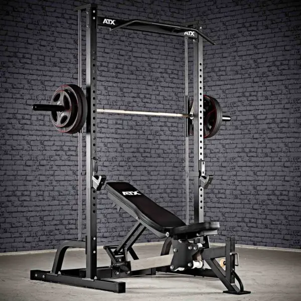 The ATX® Smith Machine Half Rack, featuring a barbell and weight plates, paired with an adjustable bench, sits against a textured gray brick wall on a concrete floor.