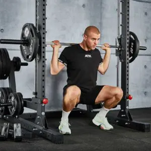 A person in a black T-shirt is performing a squat with a barbell on their shoulders at a gym. The gym equipment and weight plates are visible in the background.