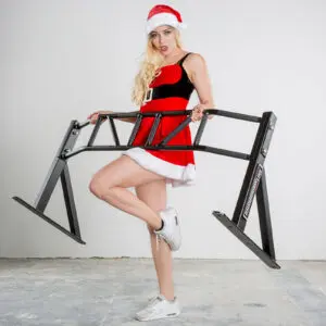 A person in a Santa hat and festive attire playfully holds the large ATX® Gladiator Chin Up Bar. They stand on a concrete floor against a plain white wall, balancing on tiptoe and wearing white sneakers.