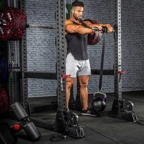 A man in a gym using the ATX® Universal Bracket during strength training with a barbell and kettlebell, stands inside a power rack, wearing a black sleeveless shirt and gray shorts amidst fitness equipment against a brick wall backdrop.