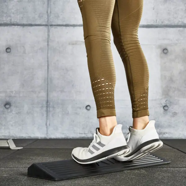 A person in olive green leggings stands on a squat wedge block performing a calf stretch, wearing white and gray athletic shoes. The background features a concrete wall and rubber flooring typical of a gym setting.