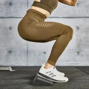 A person wearing brown leggings and white sneakers performs a squat on a squat wedge block in a gym setting. The wall in the background is made of concrete panels.