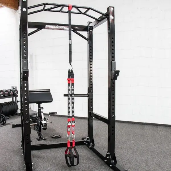 A gym setup with a black, sturdy power rack with adjustable safety bars and the ATX® Suspension Trainer Set PRO hanging from the top. An adjustable bench with black padding complements the scene, all set on a gray carpeted floor.