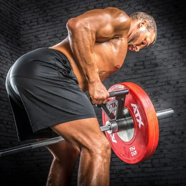 A muscular person in black shorts is performing a bent-over row with an ATX Parallel Grip T Bar Row Handle and a 7ft 1500lbs Olympic Bushing Bar, loaded with red weight plates, against a gyms gray brick wall backdrop.