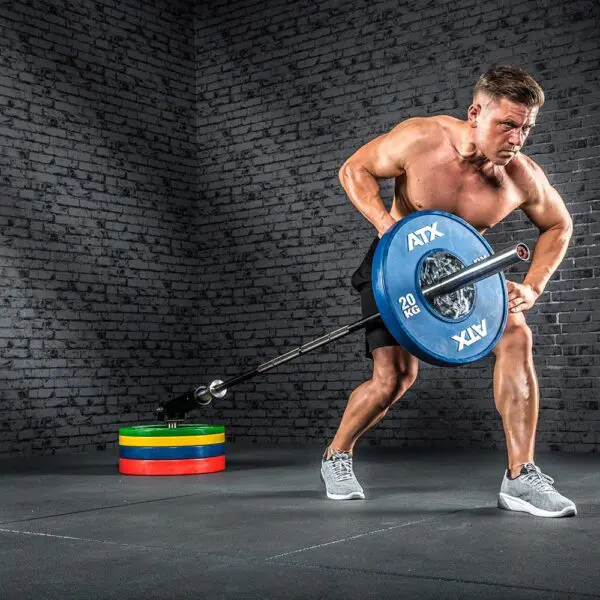 A shirtless muscular man works out in a gym, using an ATX® Single Arm Row Barbell Handle and a 20 kg weight plate. He wears black shorts and gray sneakers. Stacked colorful weight plates are behind him against a brick wall.