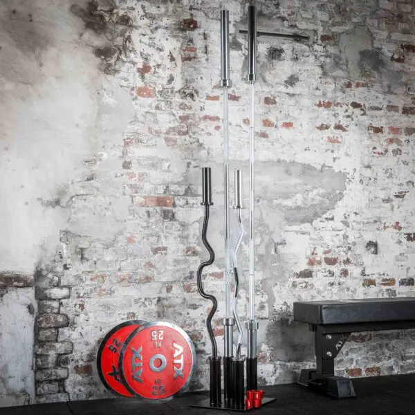 A gym setup shows an Olympic Barbell Holder with straight and curl bars against a rustic brick wall. A red 25 kg weight plate rests nearby, while a bench is partially visible on the right side.
