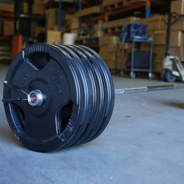 The Power Maxx 6ft Olympic Barbell, stacked with multiple 25 kg plates on both sides, sits on the gym floor. The backdrop features shelves and assorted equipment, highlighting a focused weightlifting or fitness area.