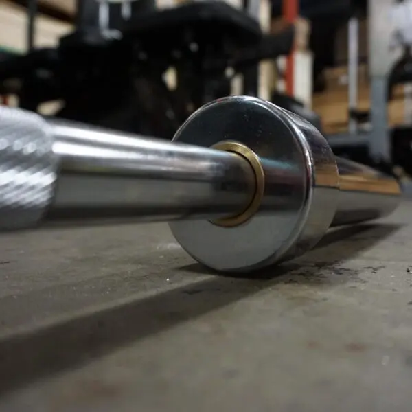 A close-up shows the 7ft Olympic Barbell Thrust Bearings 1500lbs resting on a gym floor. The shiny metal finish of the barbell end reflects the light, while blurred gym equipment and weights fill the background.