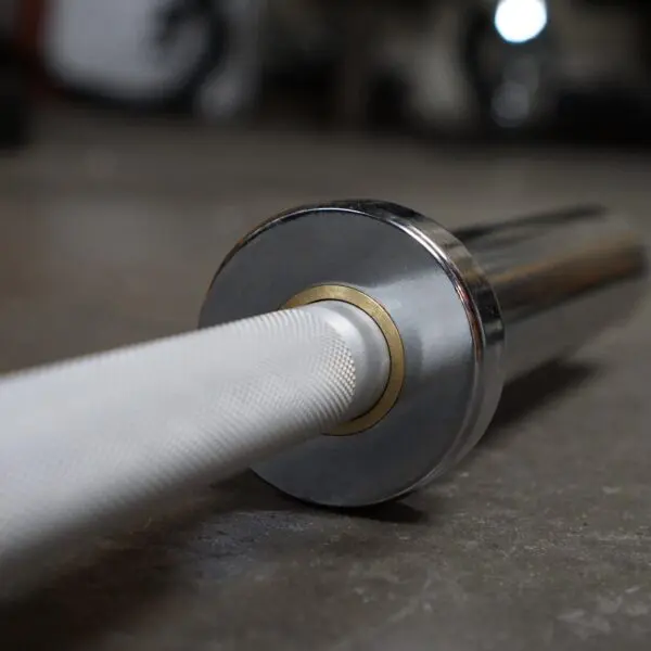 Close-up of the Aluminium 7.5kg Technique Olympic Bar resting on a gym floor. The textured grip of the metal bar contrasts with its gleaming, smooth ends. The focus is on the end part of the bar, with the background slightly blurred.