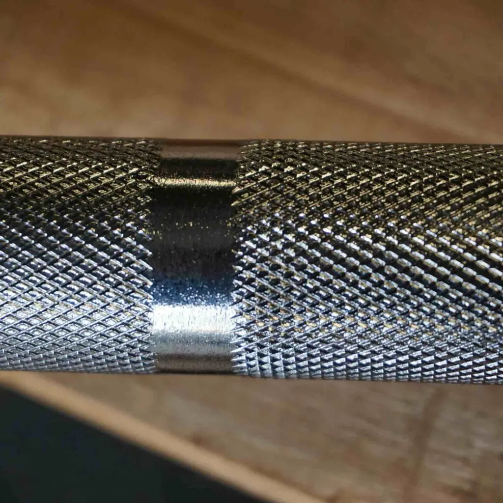 Close-up of the 7ft Olympic Barbell Thrust Bearings 1500lbs, showcasing its metallic knurled texture with a central smooth section, set against a blurred wooden background.