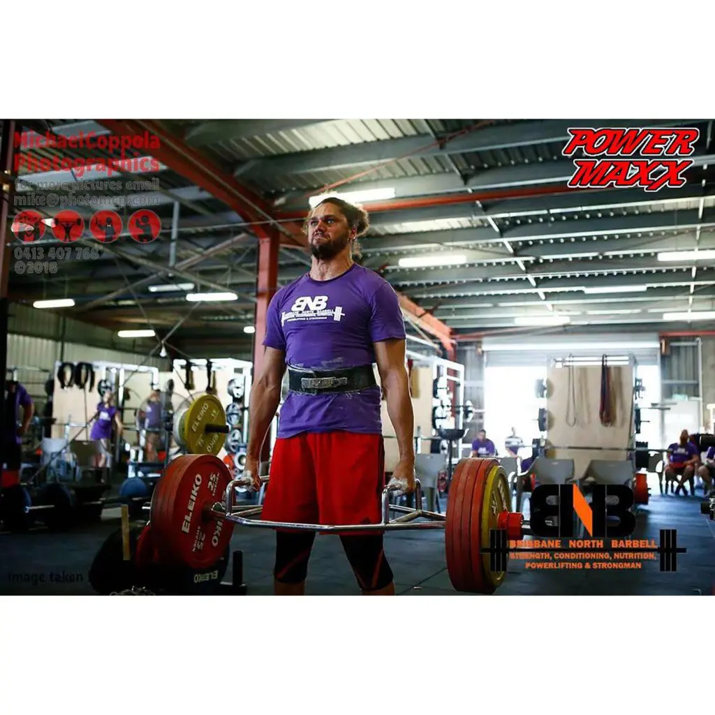 A man in a purple shirt and weightlifting belt performs a deadlift with a loaded barbell in a gym. Curious onlookers and gym equipment surround him, while tips on how to use a trap bar are featured alongside logos and contact information.