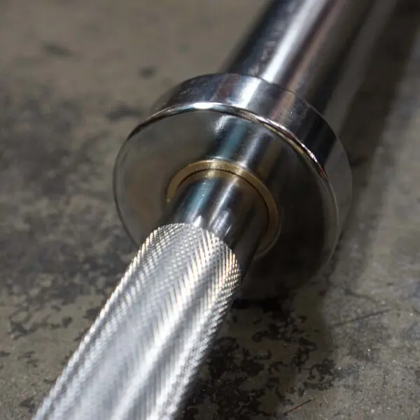 Close-up of the Power Maxx Big Olympic Dumbbell Handle on a textured concrete floor, showcasing its metallic sleeve and intricate knurled grip. The shiny finish reflects light, highlighting superior grip detailing.